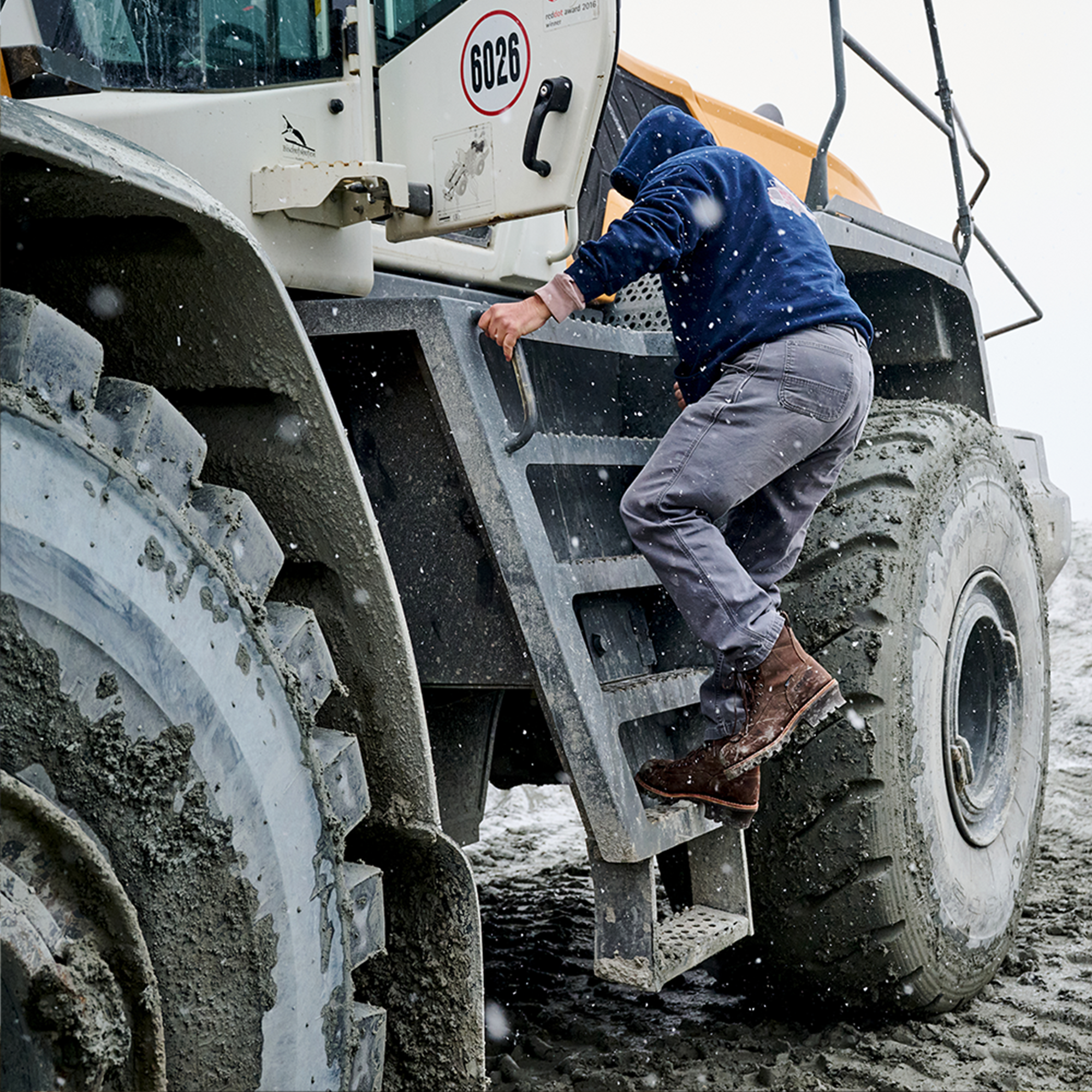 A man climbing a tractor wearing Carolina logger boots in brown.